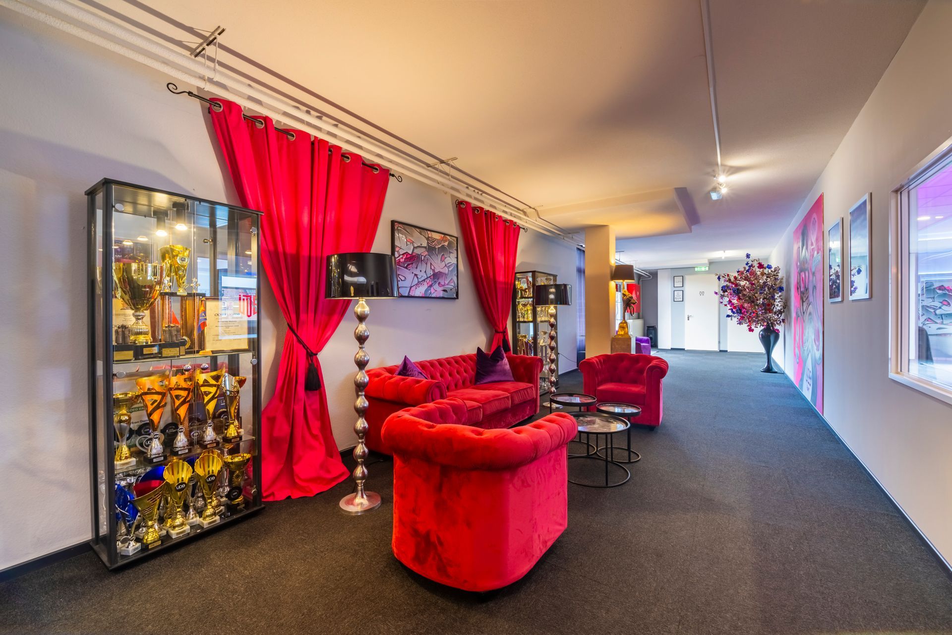 Rode lounge met fluwelen stoelen, felroze gordijnen, glazen vitrines en een lange gang met vloerbedekking.