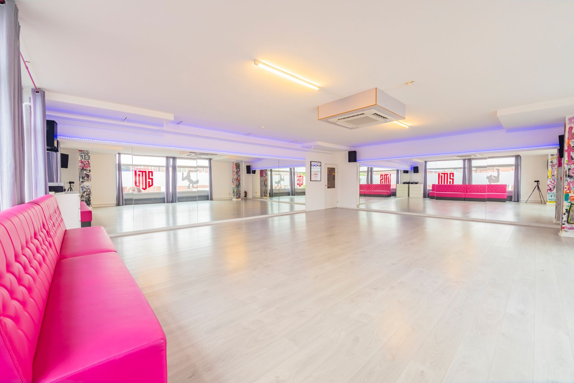 Een lichte, lege dansstudio met spiegelwanden, roze banken en grote ramen.