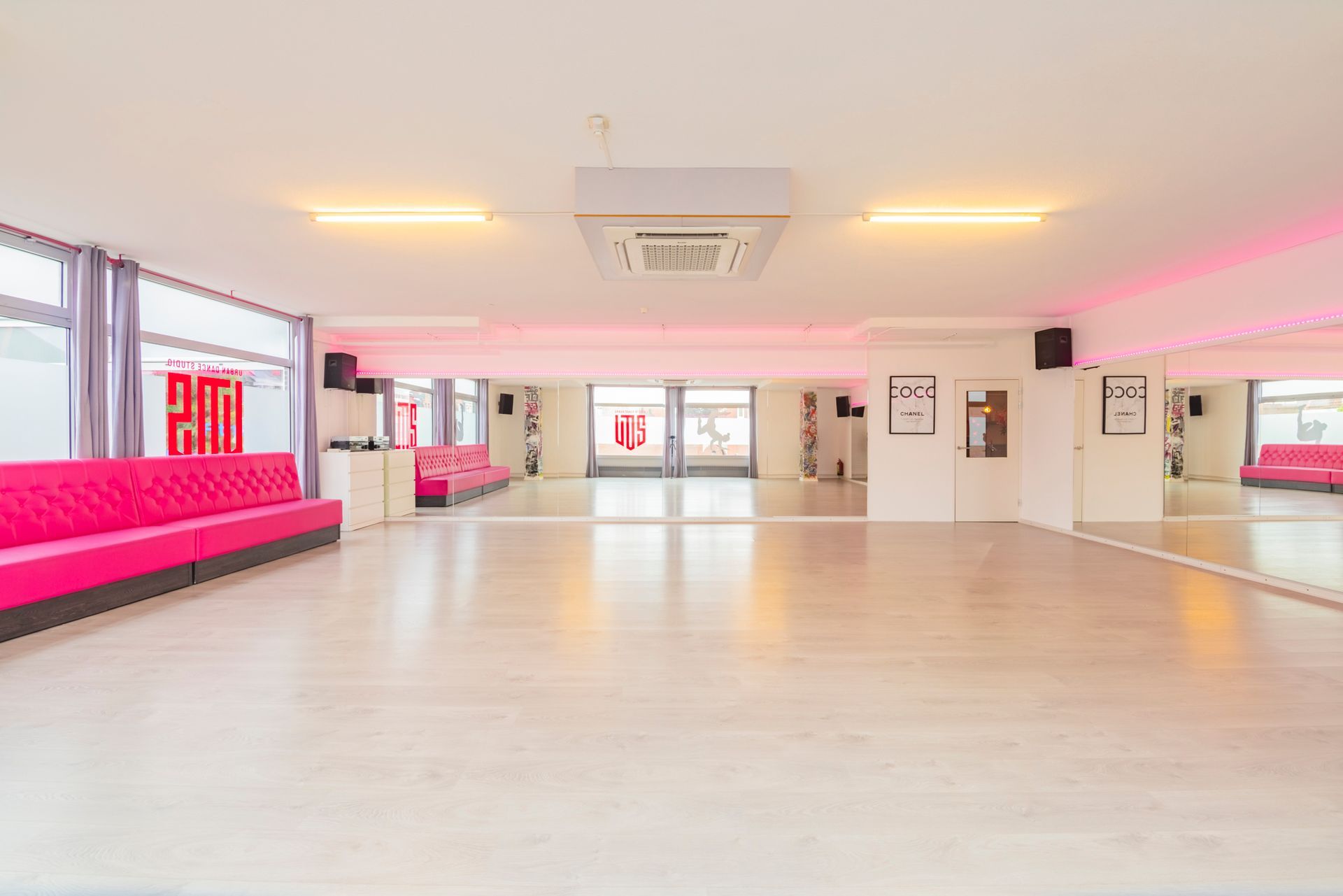 Lichte, lege dansstudio met spiegelwanden, roze banken en grote ramen.