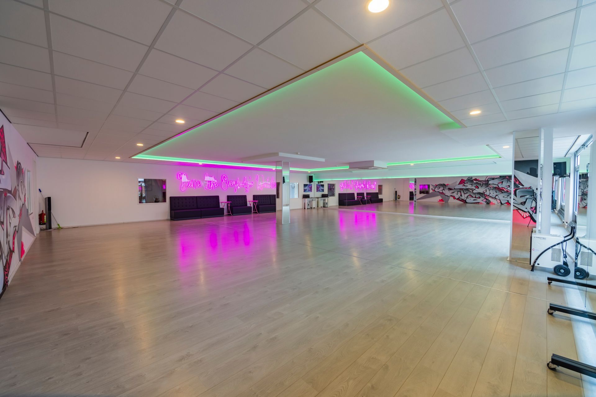 Een lege dansstudio met houten vloer, spiegelwanden en roze en groene plafondlampen.