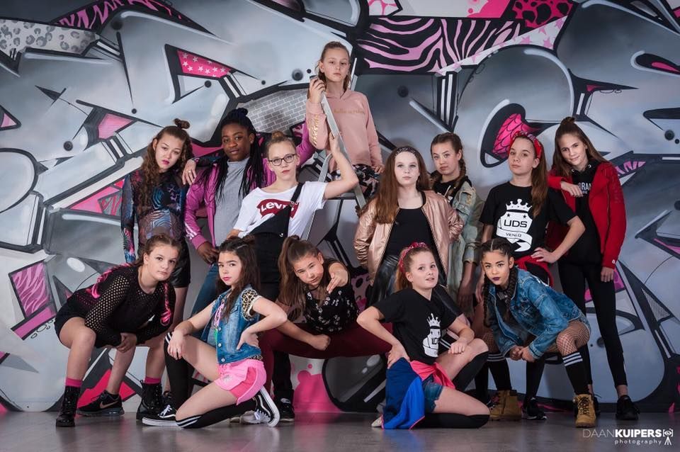 Een groep dansers poseert in kleurrijke hiphopoutfits tegen een graffitimuur.