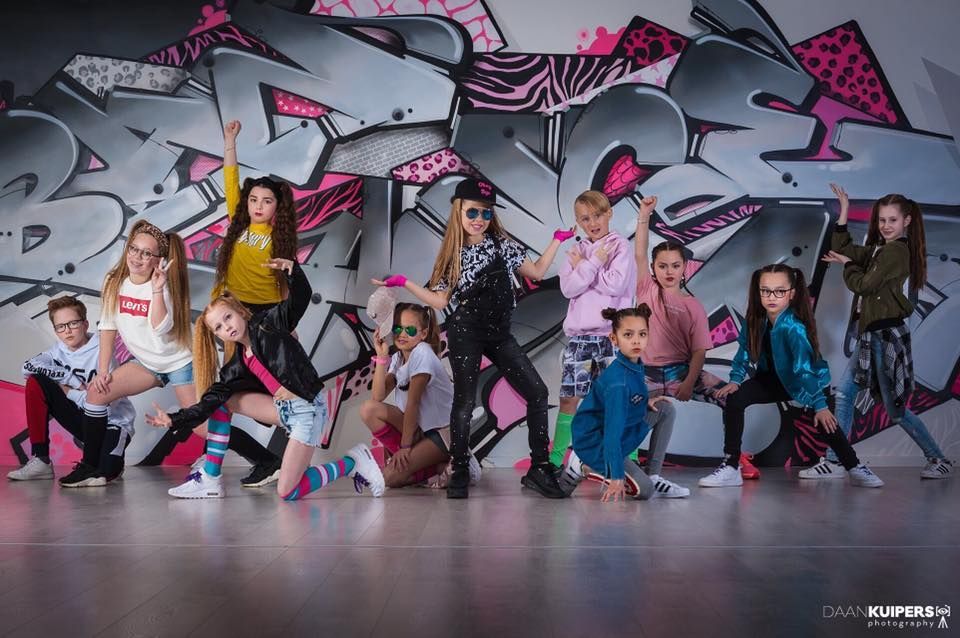 Een groep dansers poseert in kleurrijke streetwear voor een graffitimuur.
