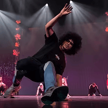 Een danseres in zwart neemt een breakdance-pose aan op een verlicht podium, met andere artiesten op de achtergrond.