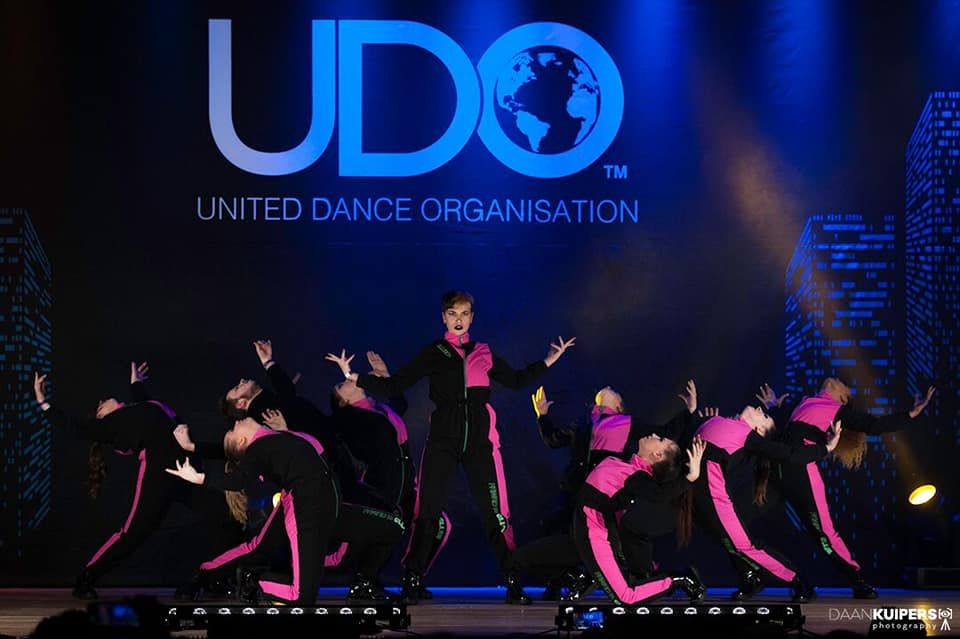 Dansgroep in zwarte en roze outfits treedt op op een podium onder een groot UDO-logo.