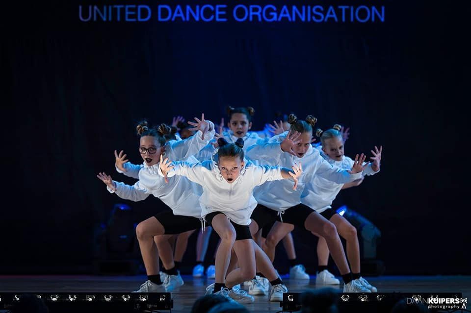 Dansgroep in witte jasjes voert een synchroondansroutine uit op een podium met blauwe verlichting.