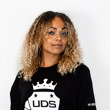 Vrouw met krullend blond haar, gekleed in een zwart UDS-shirt, tegen een witte achtergrond.