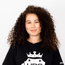 Persoon met krullend haar, gekleed in een zwart shirt, lachend tegen een witte achtergrond.