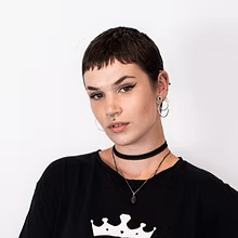 Portret van een persoon met kort donker haar, oorringen, een zwarte choker en een zwart shirt tegen een witte achtergrond.
