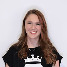 Een lachend persoon met lang bruin haar, gekleed in een zwart shirt met een witte kroonafbeelding op een effen achtergrond.
