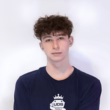 Portret van een jong persoon met krullend haar, gekleed in een donkerblauw T-shirt, tegen een effen witte achtergrond.