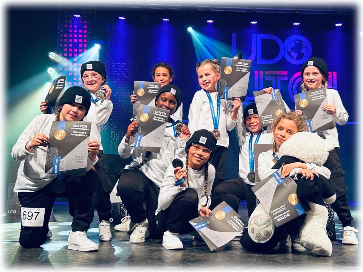 Dansgroep poseert op het podium met trofeeën en confetti, gekleed in witte outfits onder blauwpaarse lichten.