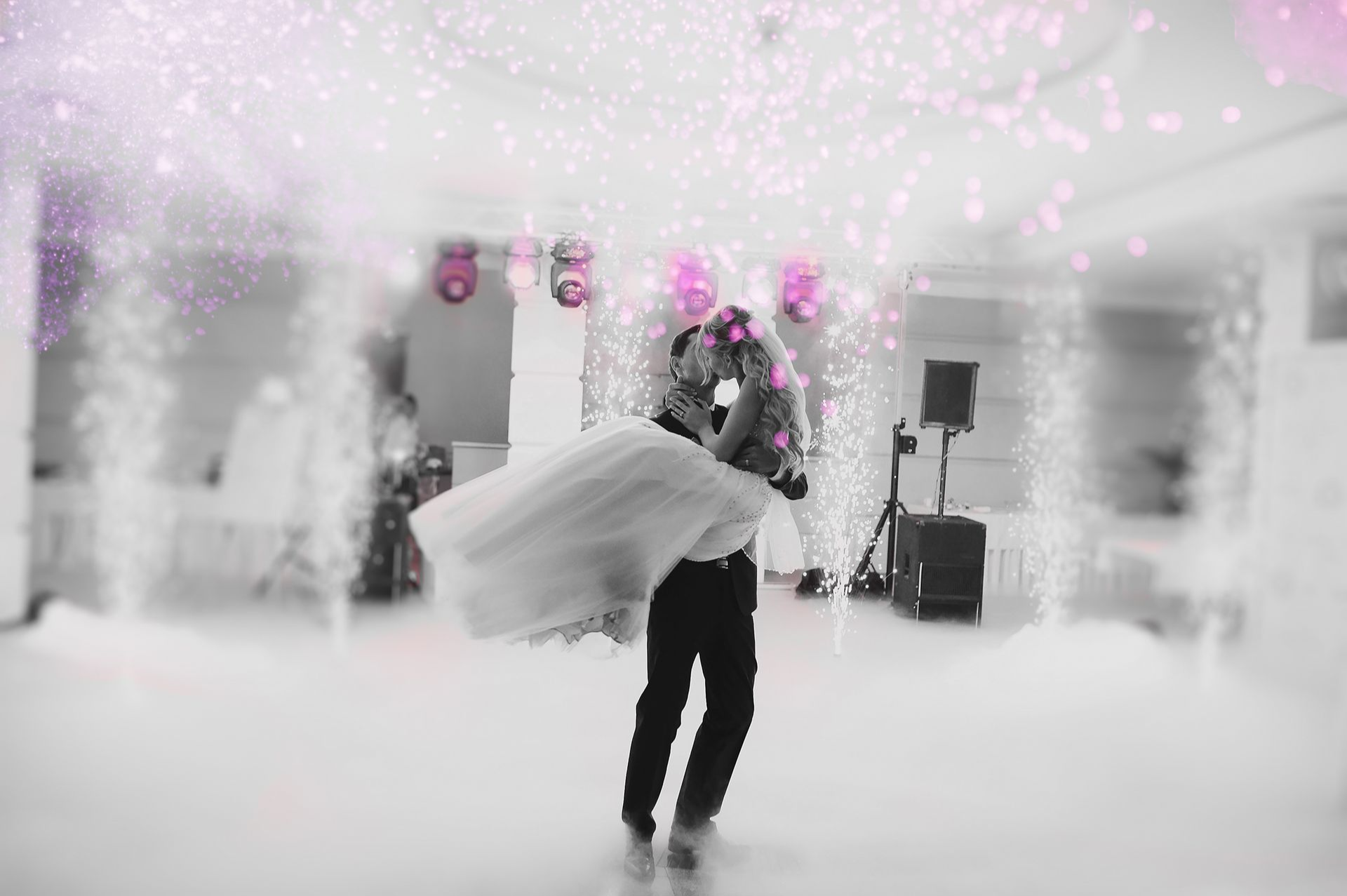 Bruid en bruidegom dansen in een witte, roze verlichte feestzaal terwijl er confetti naar beneden dwarrelt.