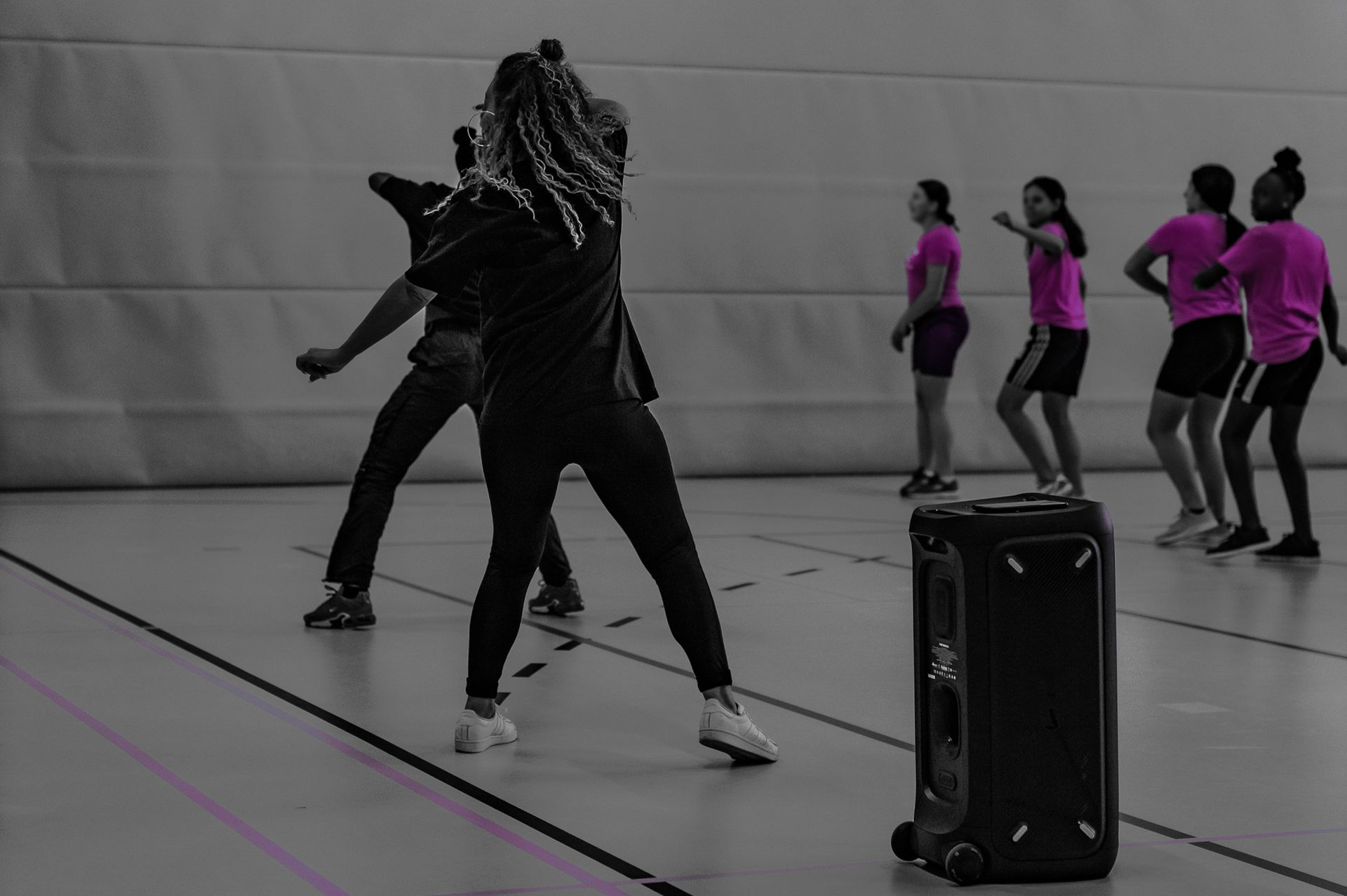 Een groep dansers in een gymzaal, met één danser in het zwart op de voorgrond en anderen in roze shirts daarachter.