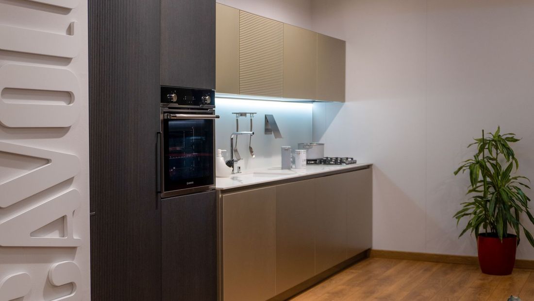 Una cucina moderna e minimalista con mobili color oro, forno nero a incasso, ripiani bianchi e una pianta in vaso.