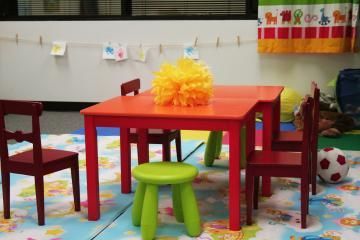 Colorful preschool play area with a red table, green stools, small chairs, and toys on a patterned rug