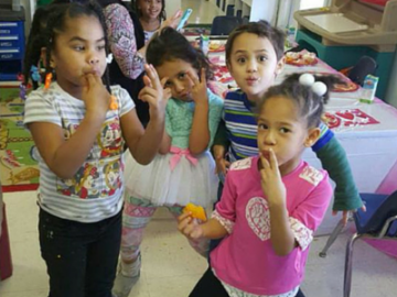 Four children standing indoors, holding small items to their mouths at a table in a classroom.