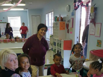 Children and adults in a classroom, with a smiling teacher near cubbies and colorful wall displays