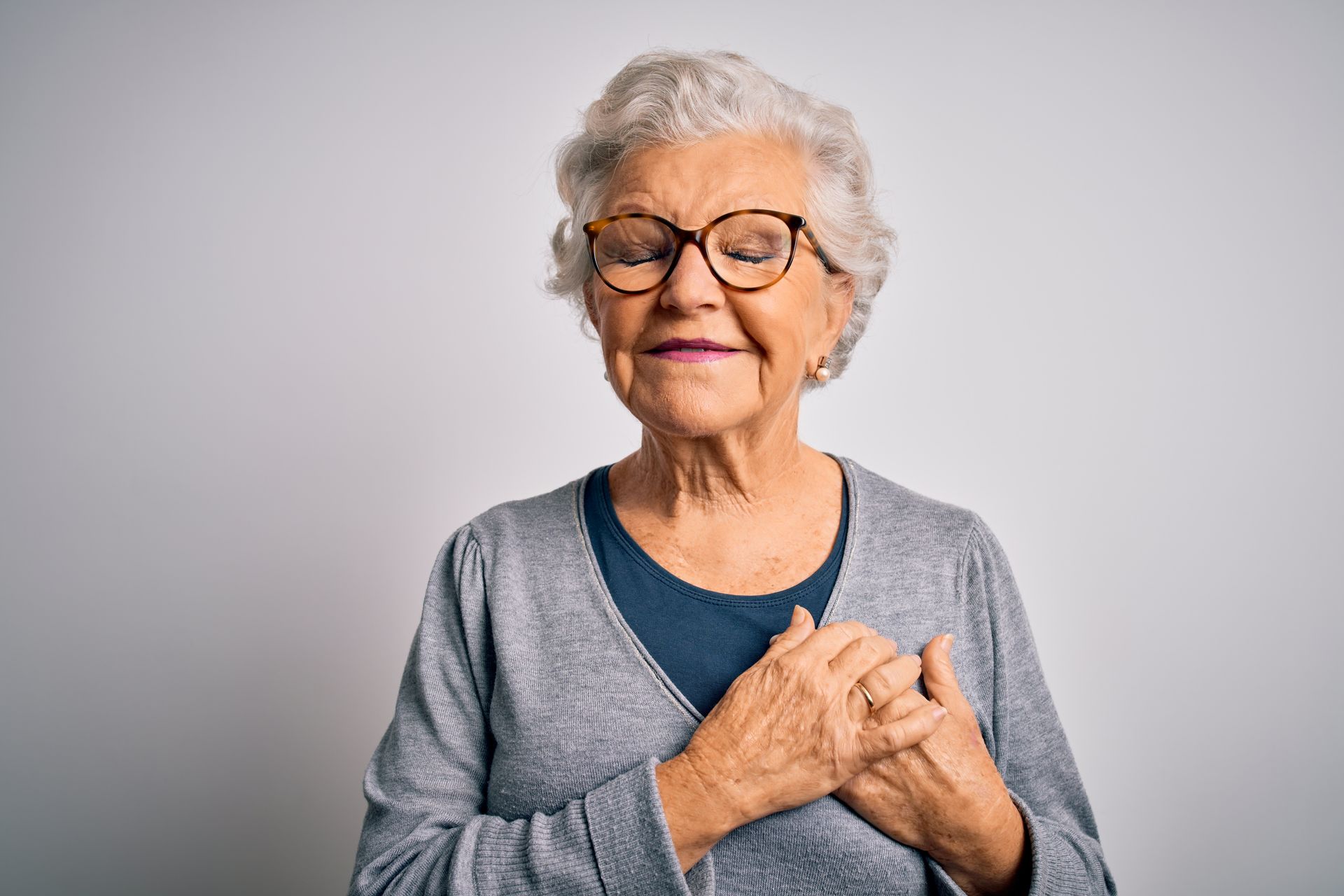 Woman wearing glasses, hands on chest, eyes closed, smiling, against a white background.