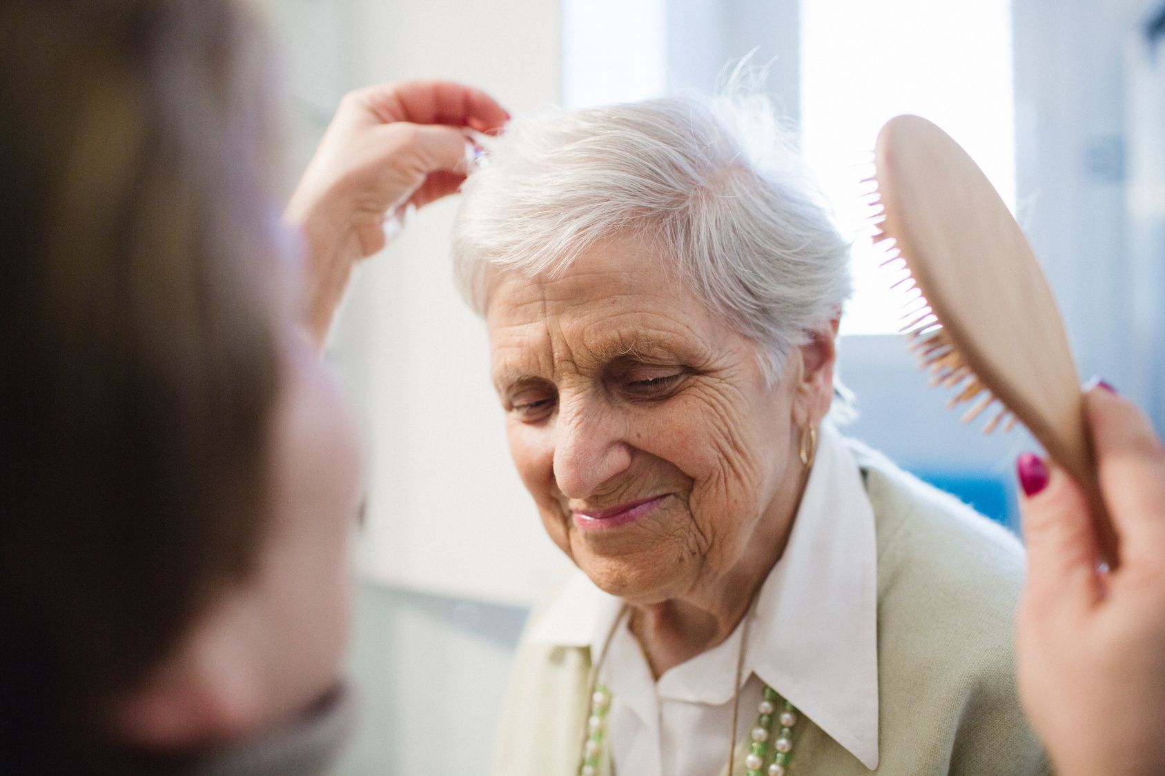 Person brushing the hair of an elderly woman. The woman has white hair and is smiling.