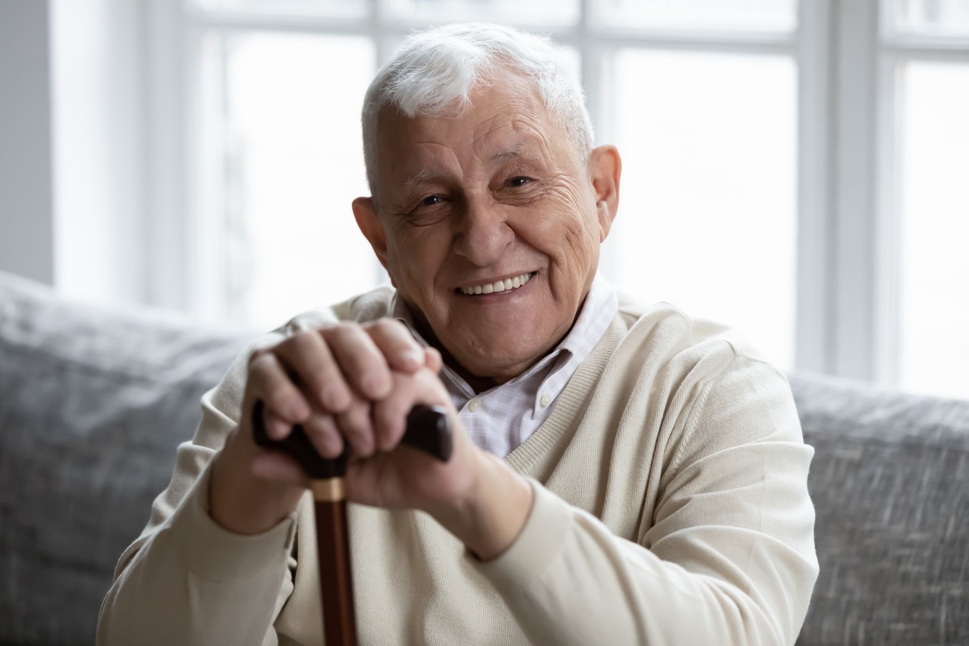 Smiling senior man with white hair, holding a cane, sitting indoors.