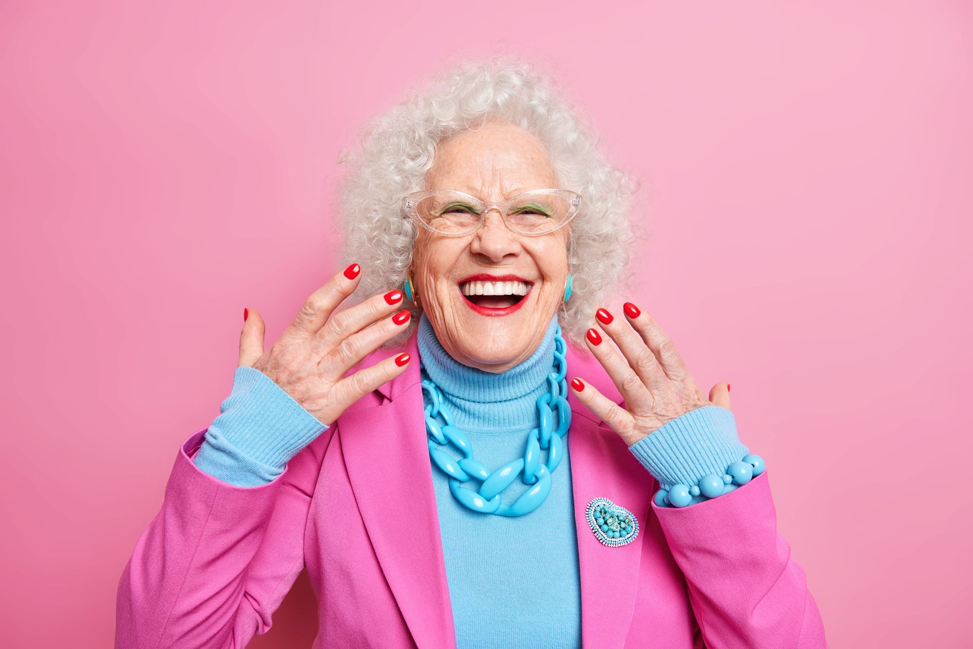 Smiling older woman with white curly hair, pink blazer, blue turtleneck and necklace, red nails, against a pink backdrop.