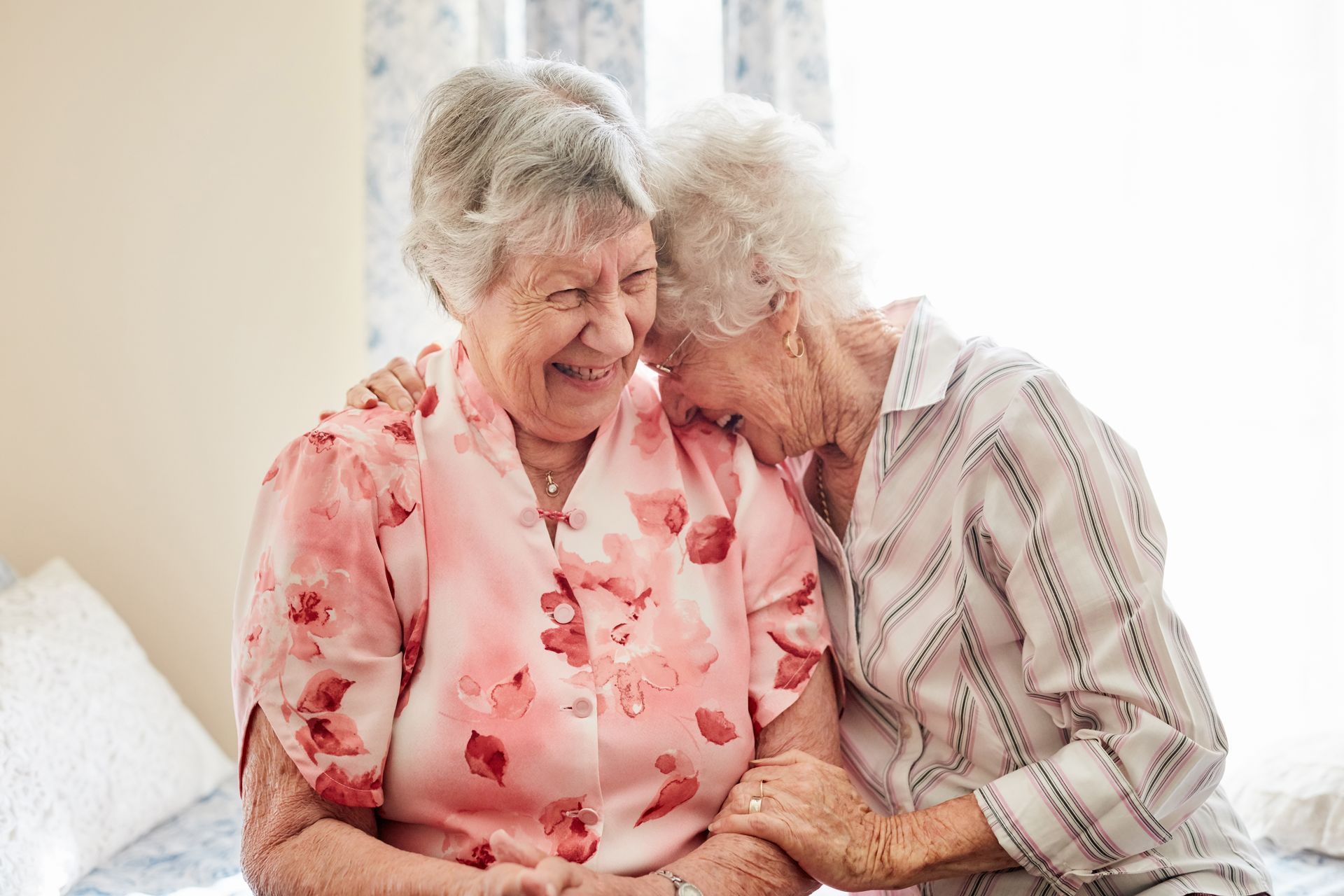 Two older women embracing, smiling, indoors. One in pink floral shirt, the other in striped shirt.