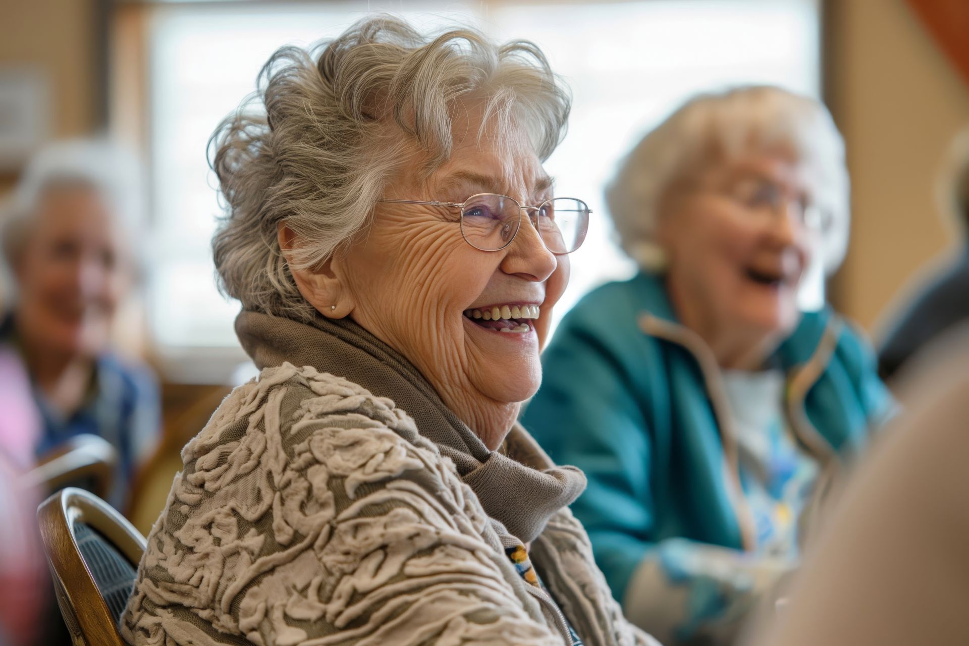 Woman with gray hair and glasses laughing, seated with other women indoors.