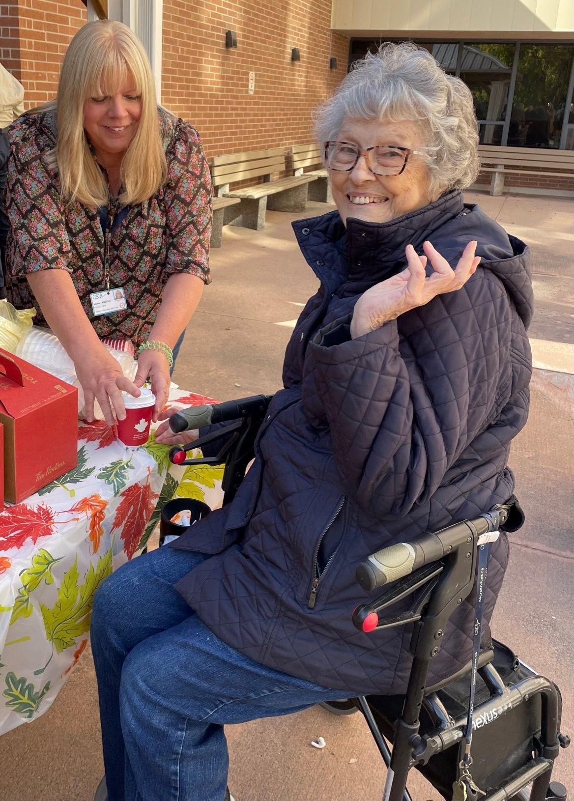 An elderly woman in a wheelchair is sitting at a table with a woman standing behind her.