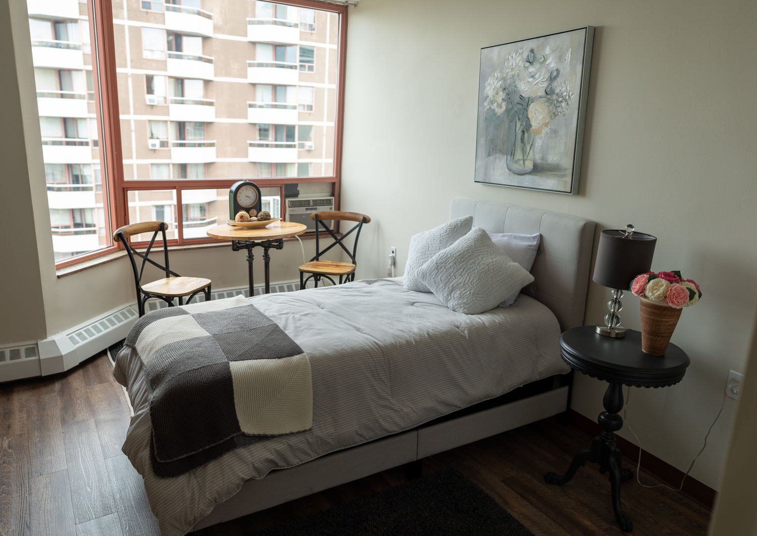 Cozy bedroom with a bed, small table, two chairs, a painting, and a window overlooking a city.