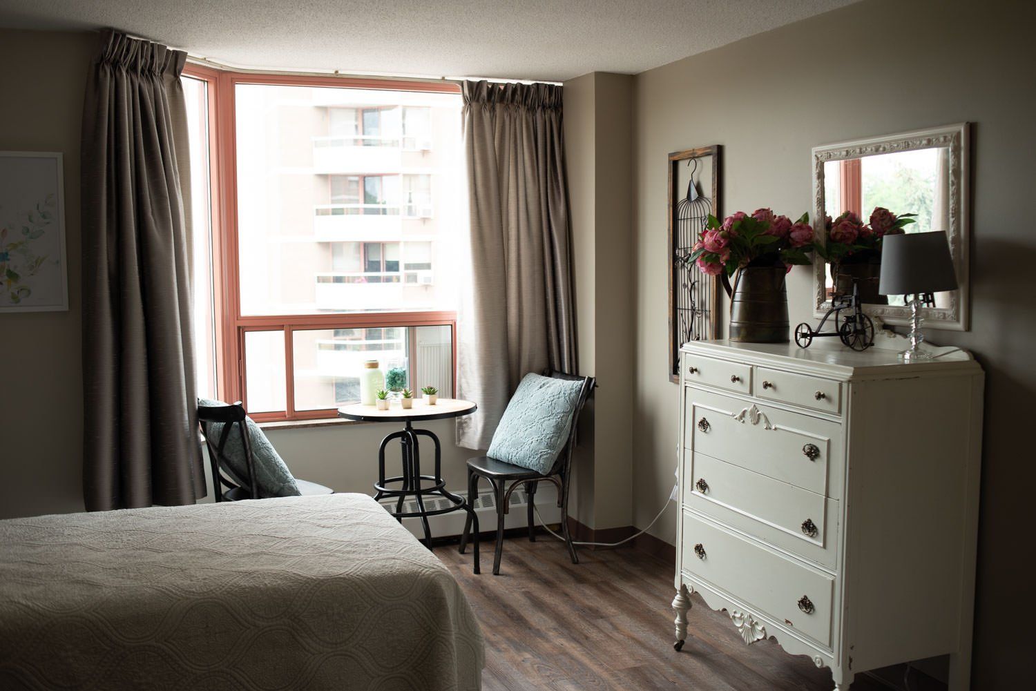 Bedroom with bed, white dresser with flowers, a round table, and curtains framing a window.