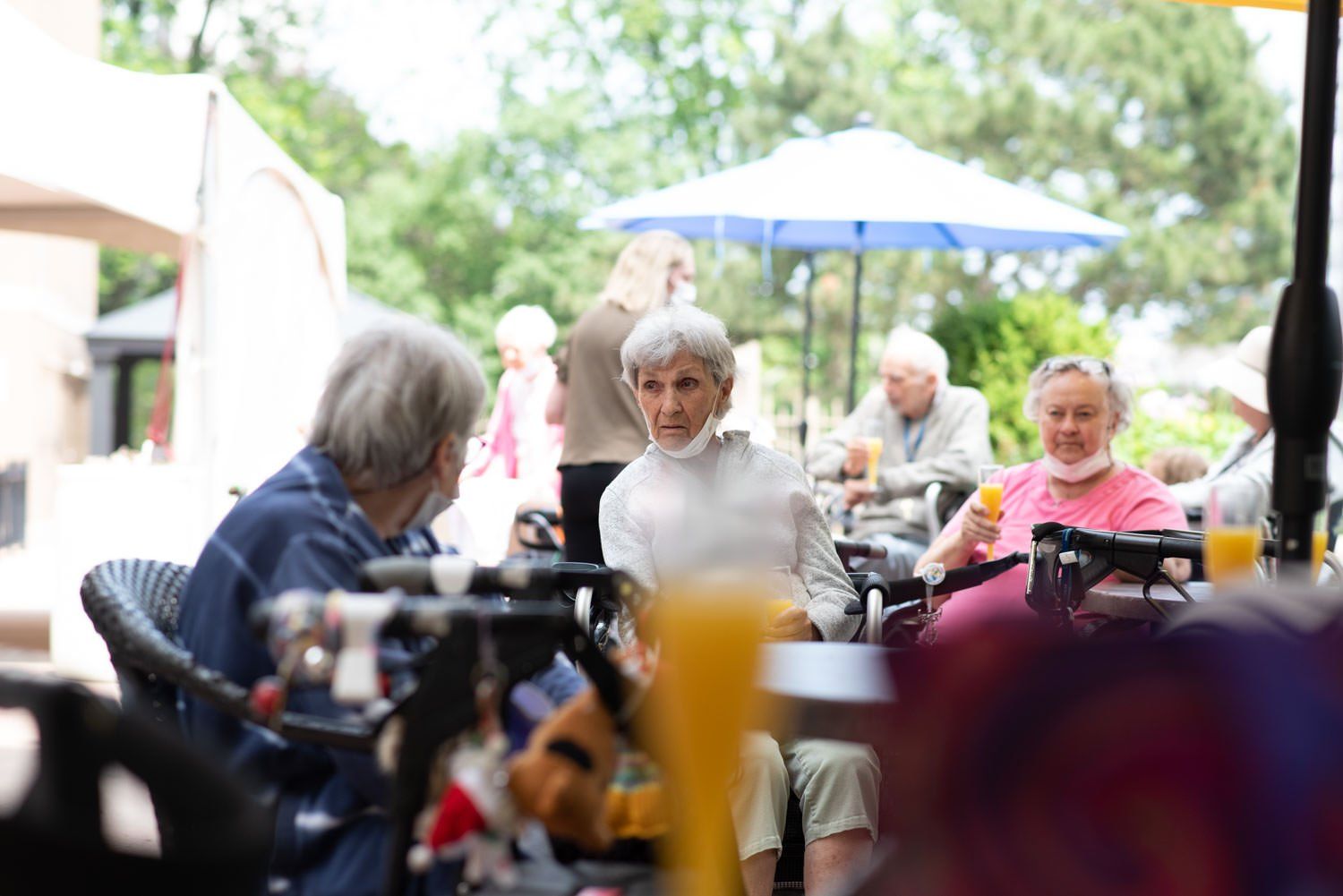 People at outdoor tables, some with walkers, drinking beverages. Green trees and umbrellas are in the background.