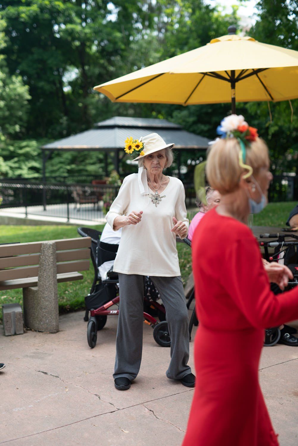 Woman dancing outside, wearing a hat with sunflowers. Others dance nearby, gazebo and yellow umbrella in background.