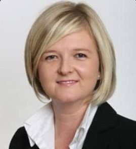 Woman with blonde bob haircut, wearing a black blazer and white collared shirt, smiling.