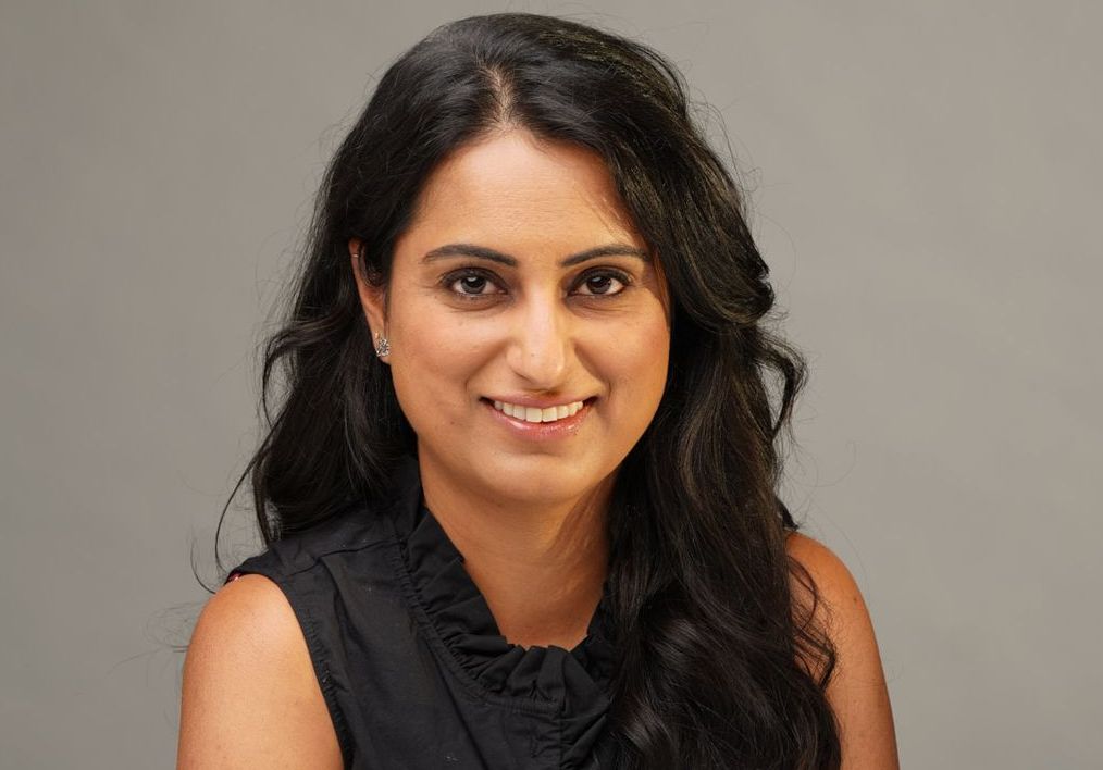 Woman with dark hair smiles, wearing a black top. Gray background.