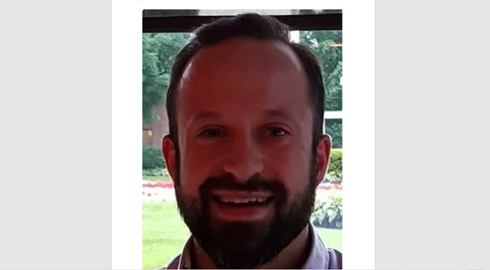 Man with a beard smiling in front of a window, looking at the camera.