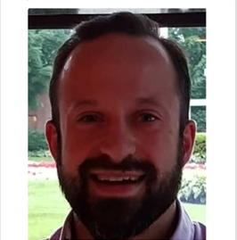 Man with a beard smiling, wearing a button-down shirt, outdoors, natural light.