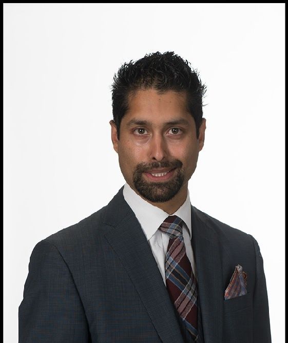 Man in a gray suit smiles, dark hair, patterned tie, pocket square. White background.