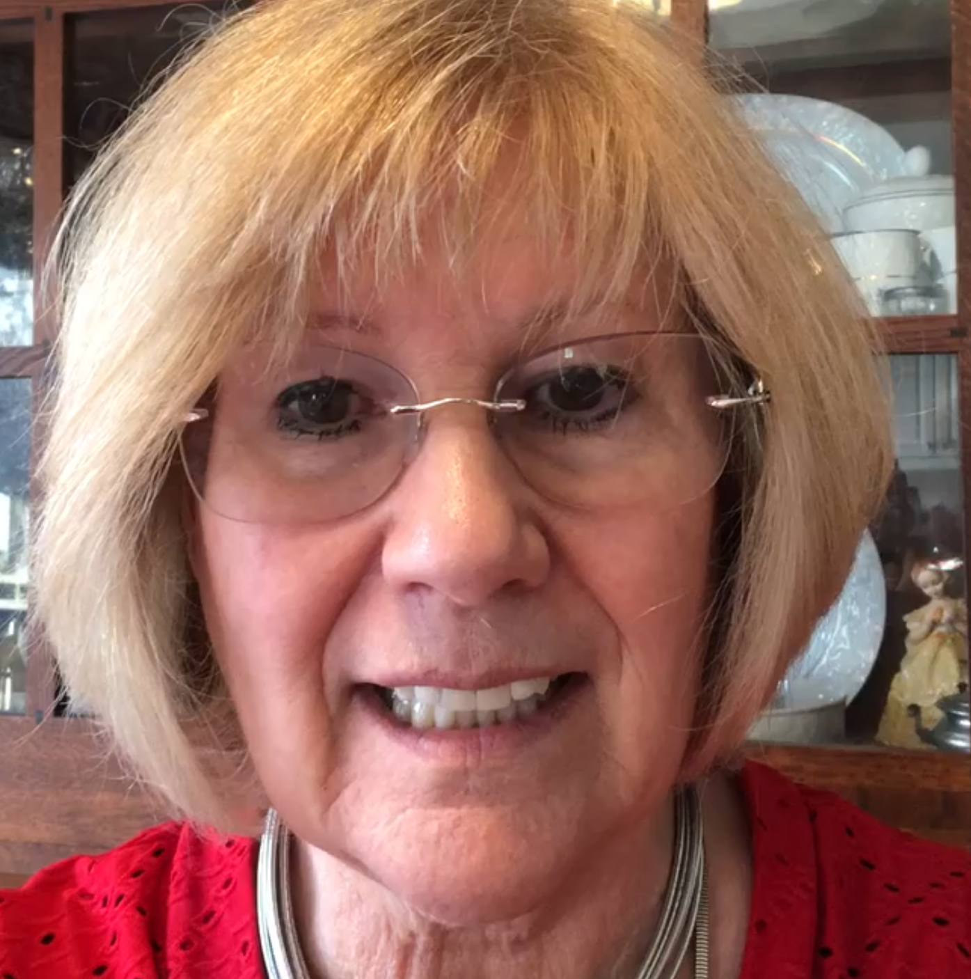 Woman with blonde bob and glasses smiles at the camera, wearing red top and silver necklace.