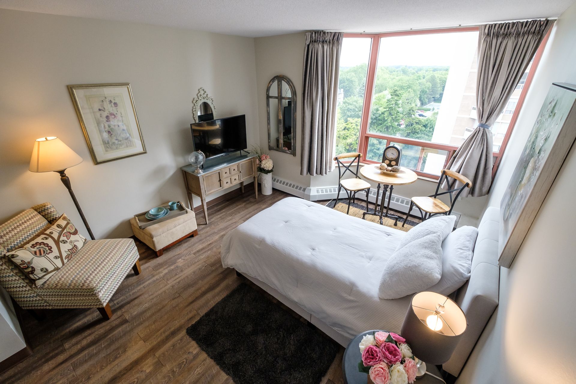 Bedroom with white bed, small table by large window, chair, and art.