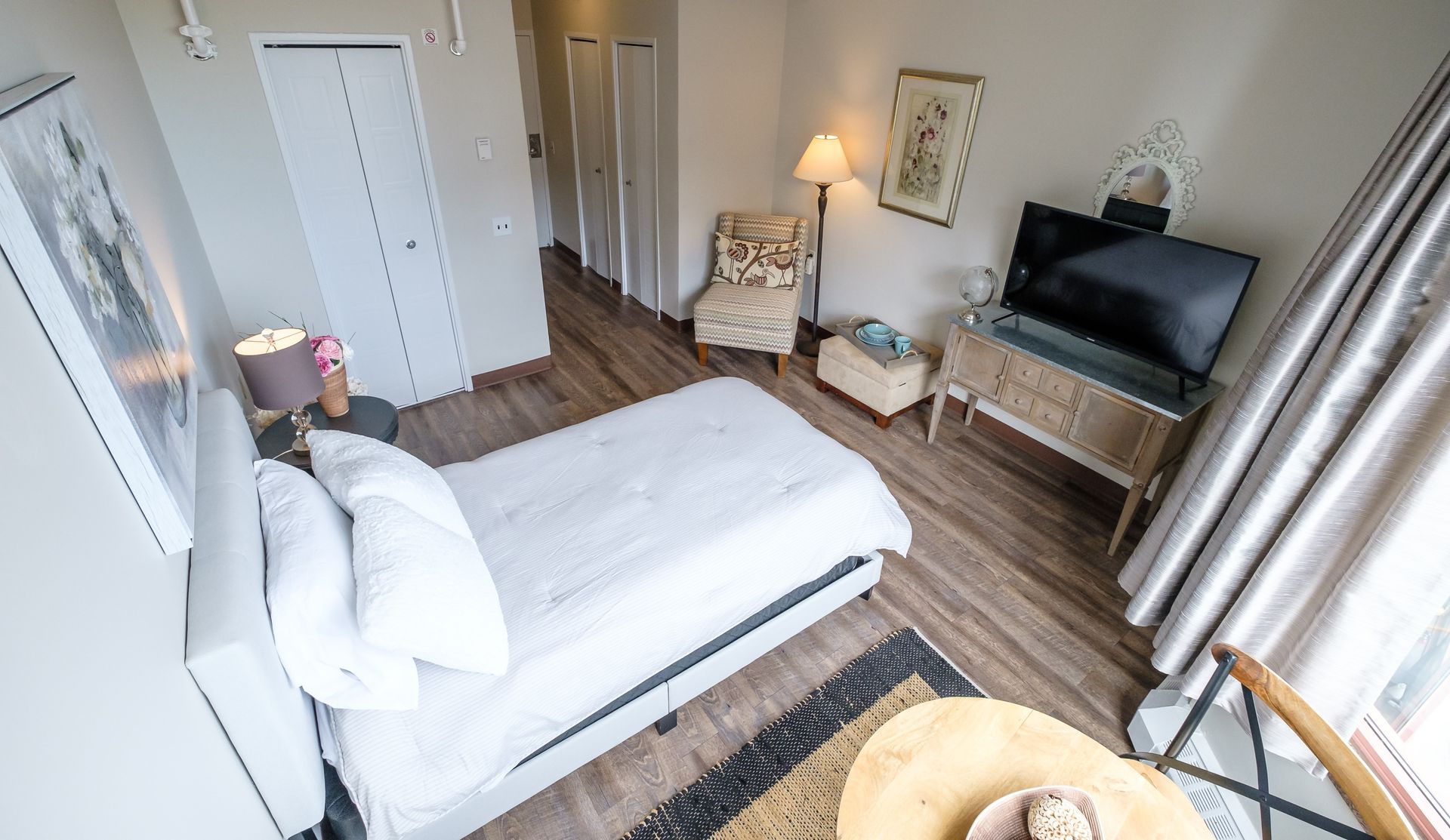 Bedroom with a bed, TV, chair, and decorative elements. Features wooden floors, a rug, and natural light.