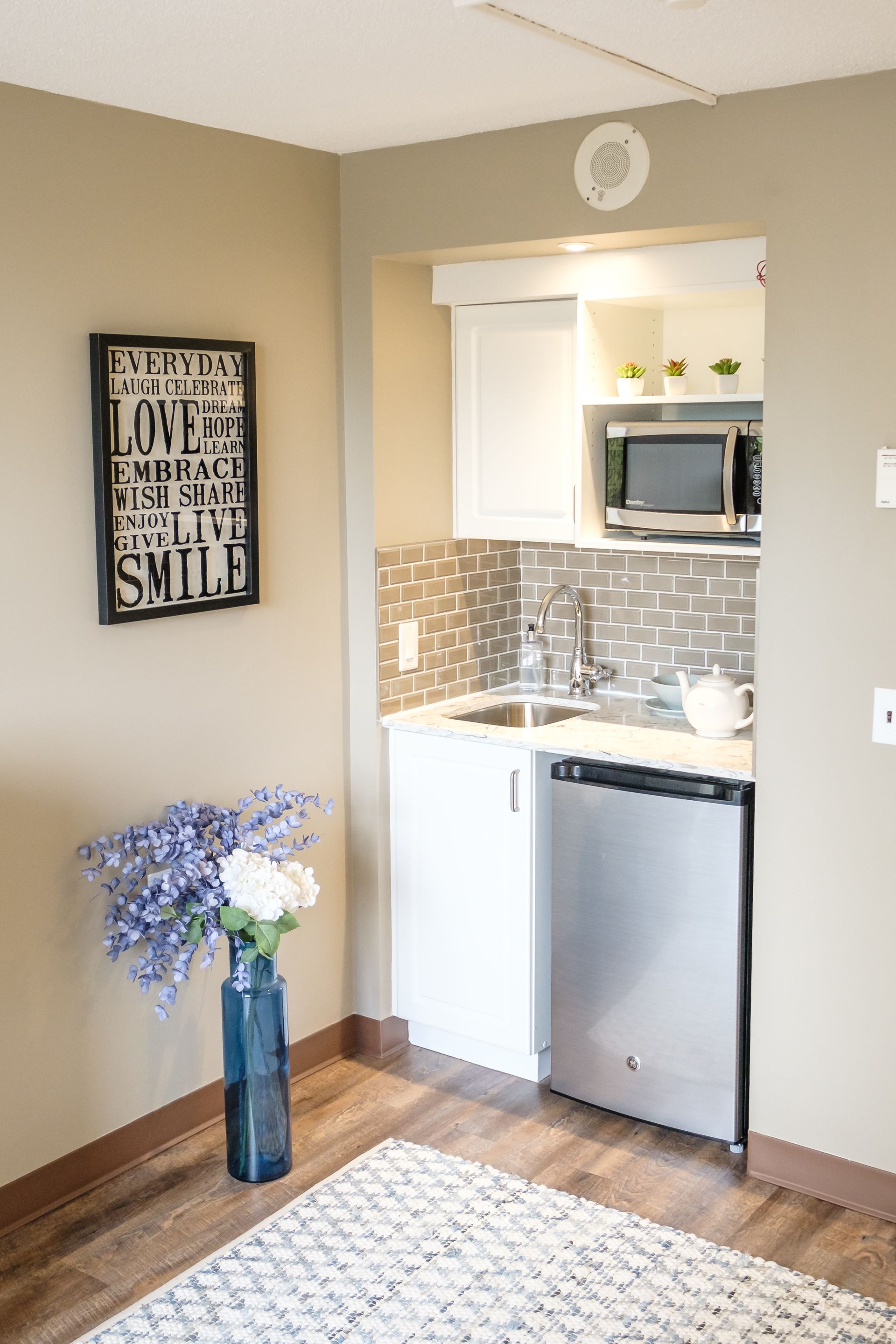 Small kitchen nook with microwave, sink, and mini-fridge. Beige walls, wood floor, decorative items.