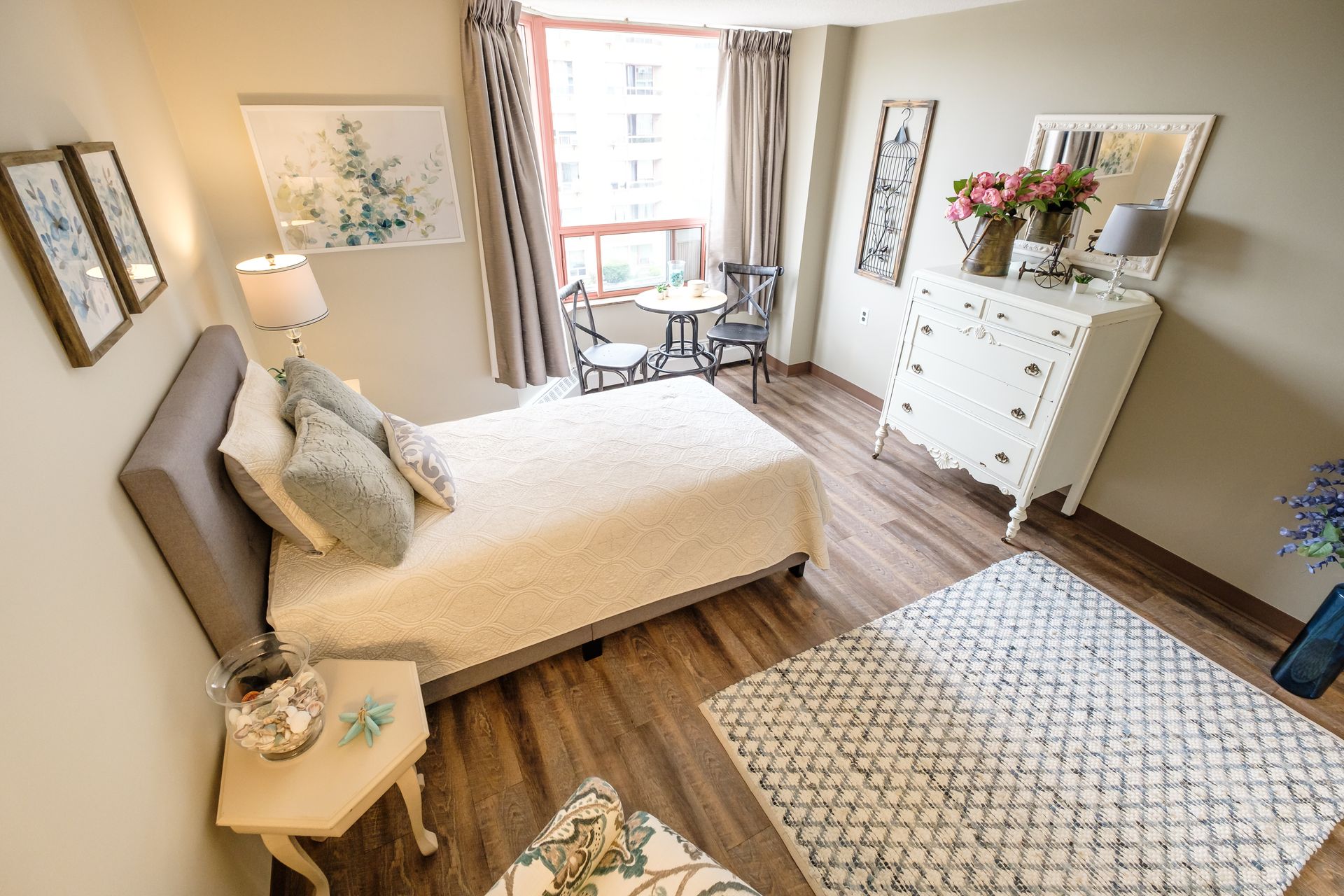 Bedroom interior with bed, dresser, table, chairs, artwork, rug, and window. Soft, neutral colors.