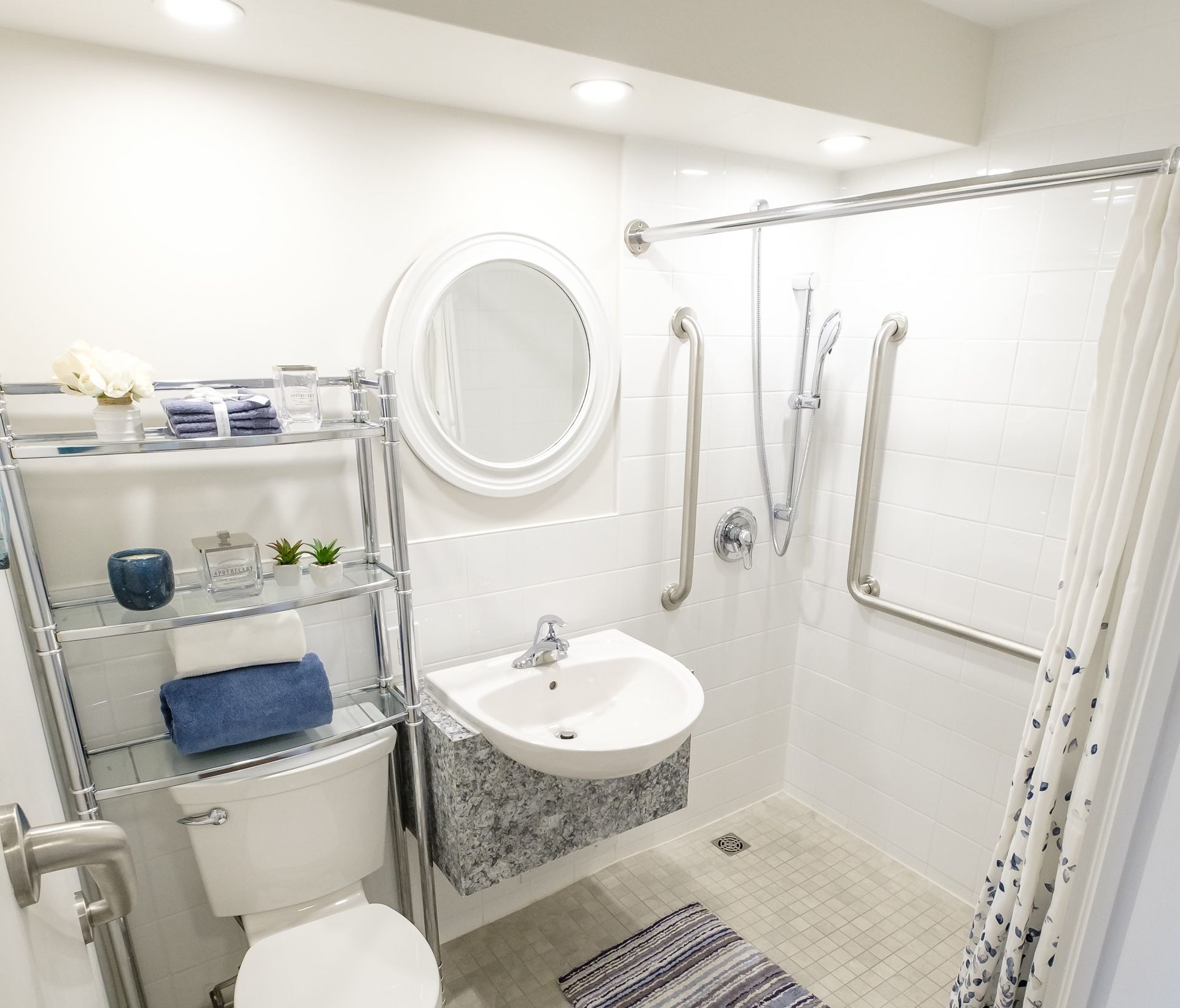 Accessible bathroom: white tiled walls, shower with grab bars, floating sink, toilet, shelving with towels.