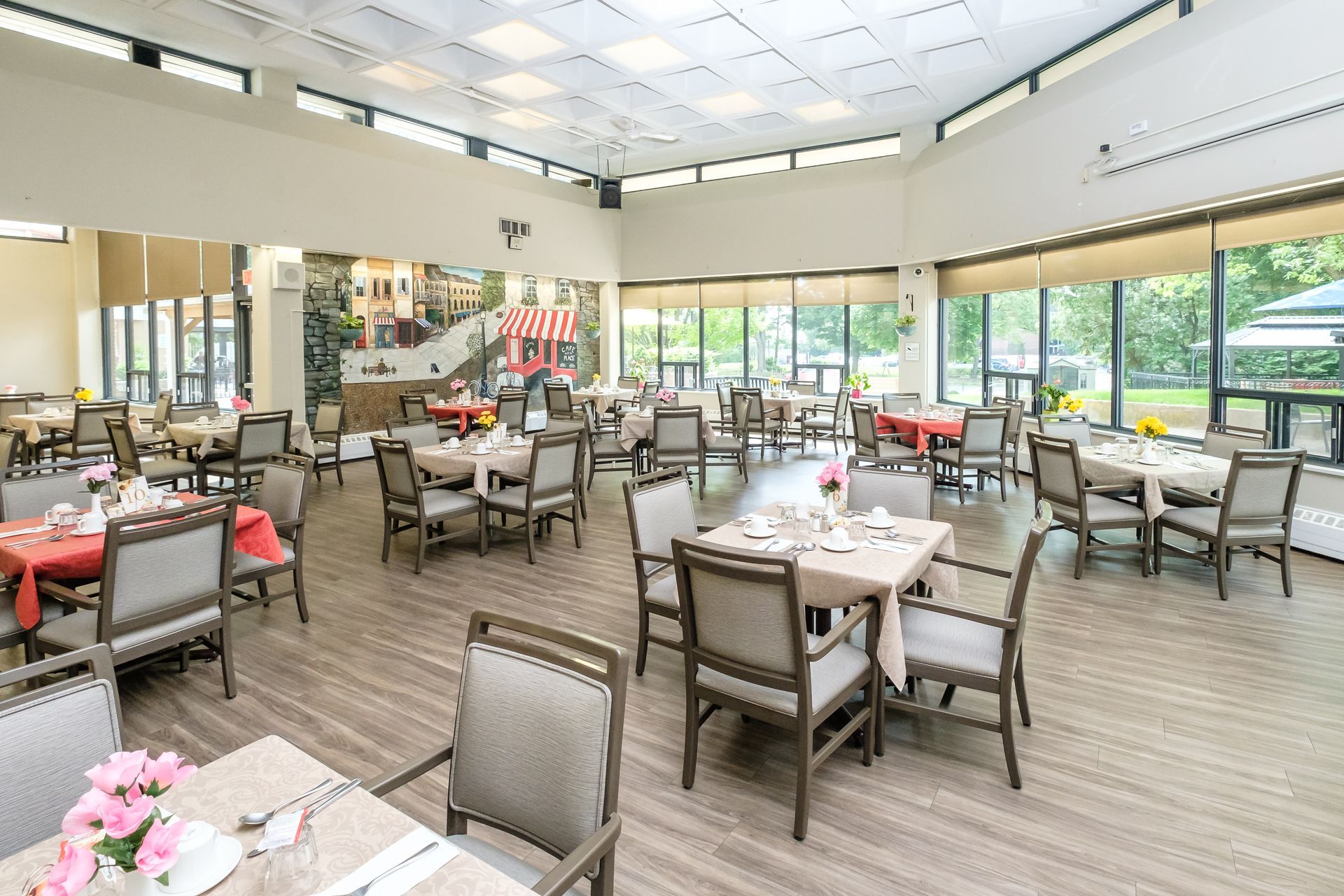 Dining room with tables set for a meal. Windows let in natural light, beige chairs, and colorful tablecloths.