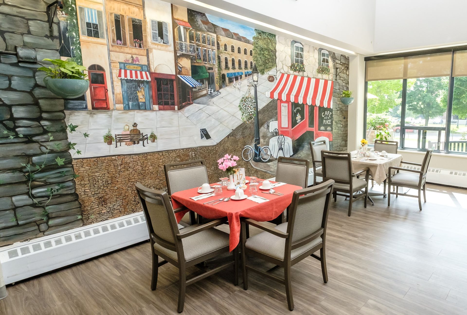 Dining room with mural of a European street scene; tables set for meals, sunlight through a window.