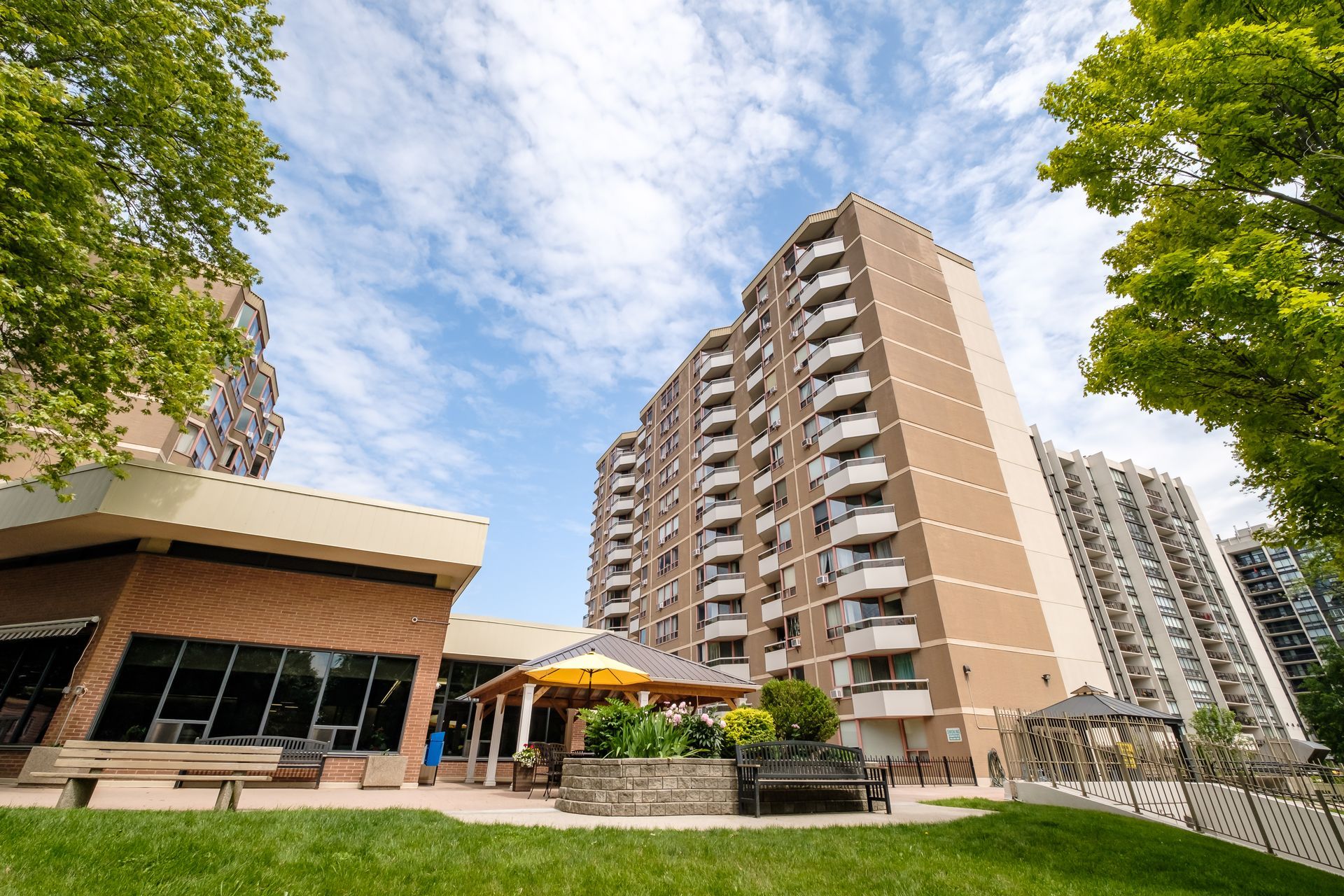 Exterior view of a tall brick apartment building with balconies, a gazebo, and green lawn under a blue sky.
