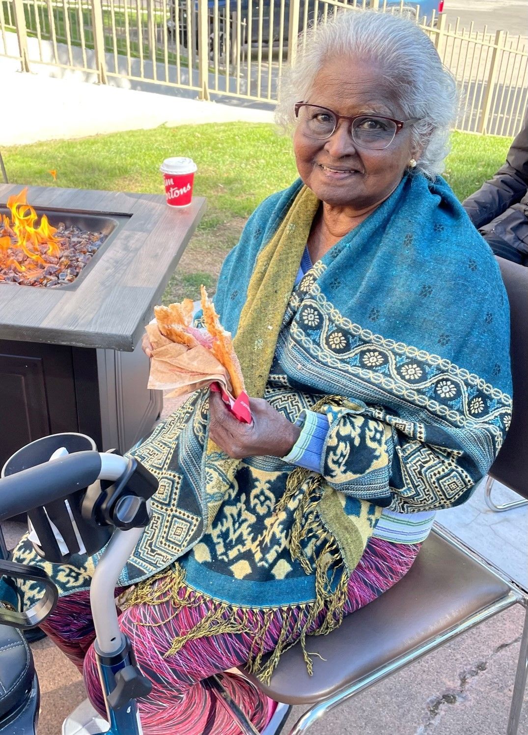 An elderly woman is sitting in front of a fire pit eating a sandwich.