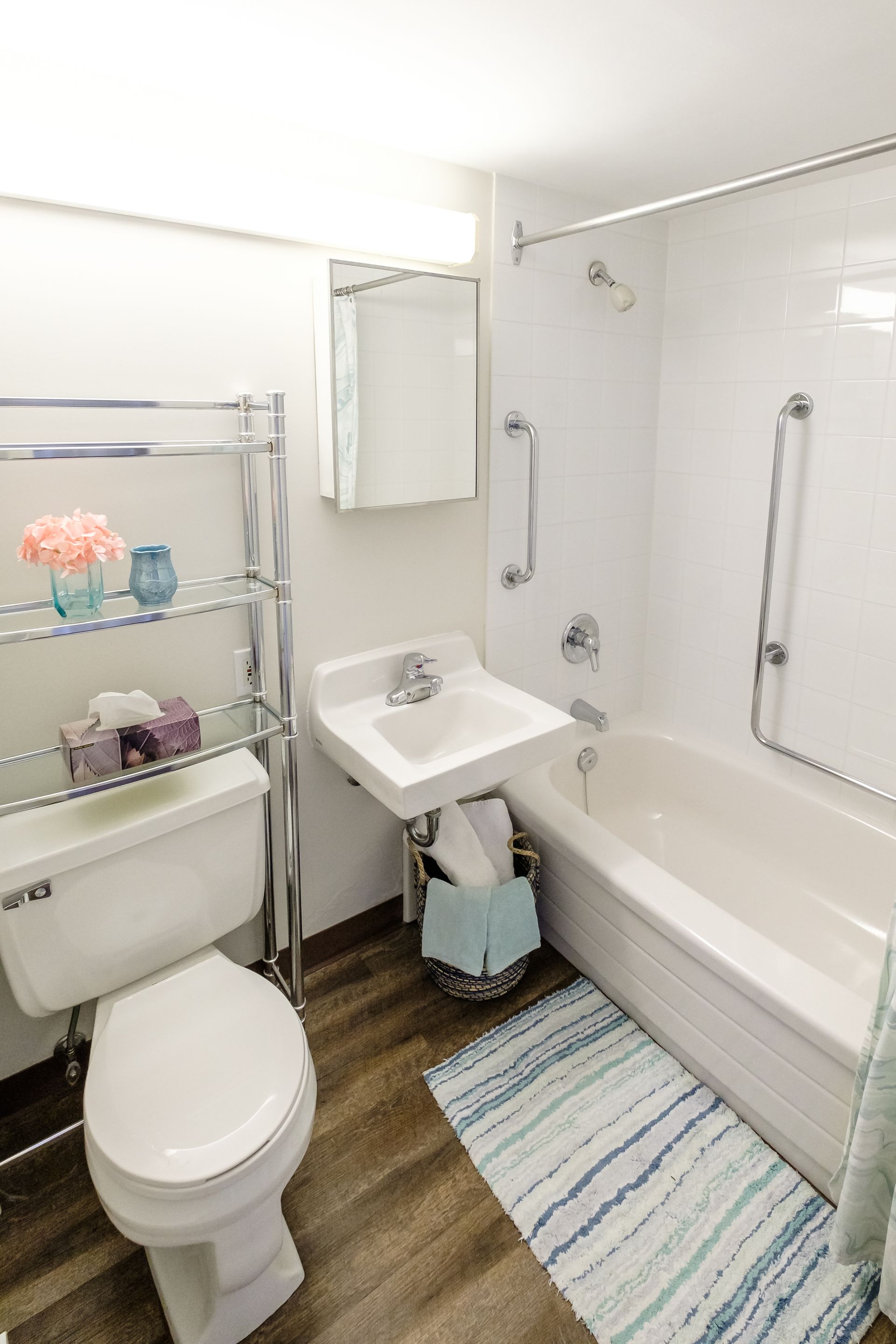White bathroom with tub, toilet, and sink. Chrome shelving, blue rug, and wood-look flooring.