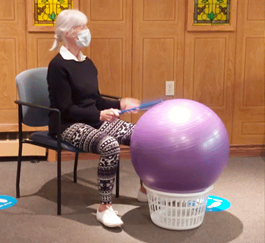 Woman in mask seated, holding sticks, hitting exercise ball on a basket. Indoors.