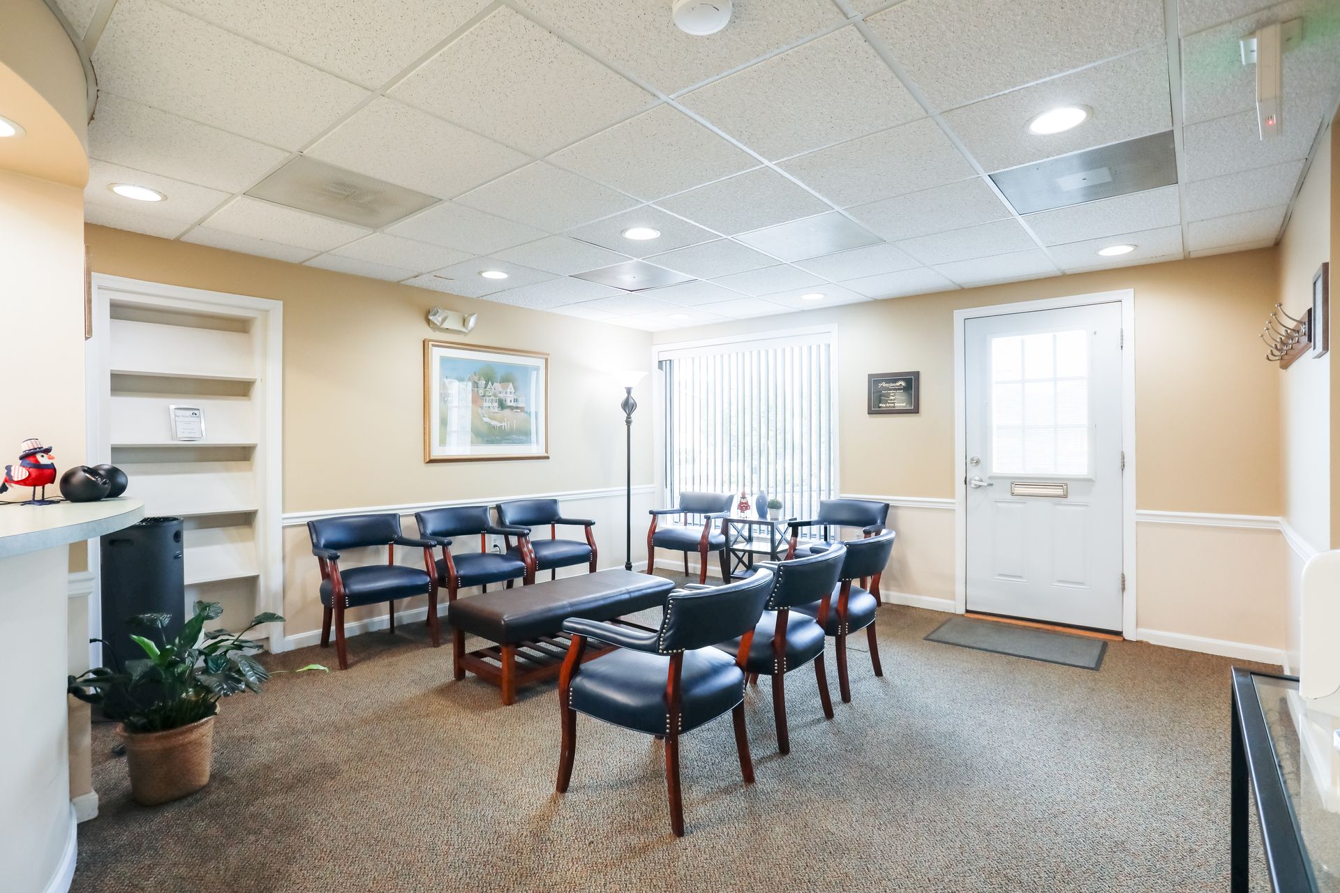 Bright waiting room with blue chairs, tables, potted plants, and a white door on the right.